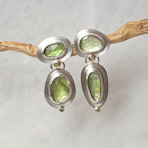 May include: A pair of silver earrings with a brushed metal finish. Each earring features a light green, irregularly shaped gemstone set within an oval frame. Small gold accents and a small green gemstone are also present.