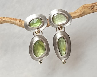 May include: A pair of silver earrings with a brushed metal finish. Each earring features a light green, irregularly shaped gemstone set within an oval frame. Small gold accents and a small green gemstone are also present.
