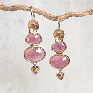 Könnte beinhalten: Ein Paar baumelnder Ohrringe mit Gold- und Silberdesign. Jeder Ohrring hat drei rosa, ovale Steine mit goldenen Rändern. Die Ohrringe hängen an einem silbernen Haken und werden auf einem Ast präsentiert. Die Steine sind leuchtend rosa.