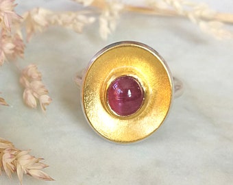 May include: A gold-plated ring with a silver band features a central, round, deep pink gemstone. The gold setting has a textured, brushed appearance. The ring is set against a soft, neutral background with dried floral accents.