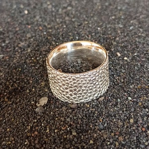 May include: A silver ring with a textured, honeycomb-like pattern. The ring is wide and has a polished interior. The ring is displayed on a dark, textured surface.
