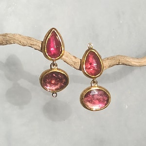 May include: A pair of gold-toned drop earrings. Each earring features a teardrop-shaped stone at the top and an oval-shaped stone below, both in a vibrant pink hue. The stones are set in gold bezels, creating a striking contrast. A small, clear stone accents each earring.