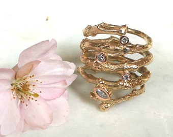 May include: A collection of gold-coloured rings with a branch-like design, featuring small, clear gemstones. A delicate pink flower is positioned to the left of the rings. The rings have a textured surface and a unique, organic aesthetic.
