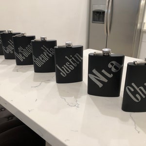 May include: Six black metal hip flasks with silver caps and personalized names in silver lettering. The names are:  'Nath', 'Lynn', 'De'Shona', 'Shenetta', 'Justin', 'Mia', and 'Chris'.