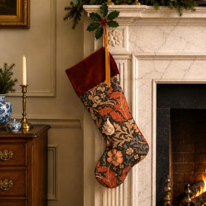 May include: A Christmas stocking featuring a deep red velvet cuff and a hanging loop. The body of the stocking displays a dense botanical pattern with orange, cream, and muted green floral and leaf motifs set against a dark charcoal background.