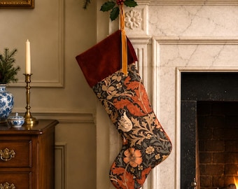 Puede incluir: Un calcetín navideño con un puño de terciopelo rojo oscuro y un lazo para colgar. El cuerpo del calcetín muestra un denso patrón botánico con motivos florales y de hojas en naranja, crema y verde apagado sobre un fondo gris oscuro.