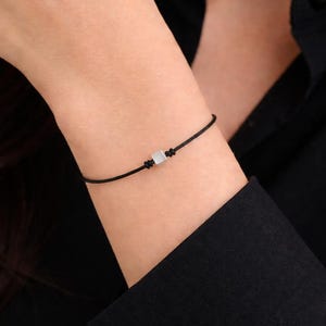 Op de afbeelding: Een minimalistische zwarte armband met een kleine, vierkante, lichtgekleurde kraal in het midden. De armband is gemaakt van een dun, zwart koord en wordt om een pols gedragen, tegen een zwart overhemd.