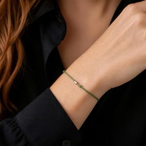 Può includere: Un bracciale sottile in cordino verde oliva caratterizzato da una piccola perlina metallica rettangolare color argento centrata sul filo. Il bracciale è realizzato in un materiale liscio e semplice e include un piccolo nodo vicino al dettaglio.