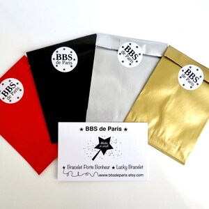 May include: Four small gift bags in red, black, silver, and gold with white circular labels that say "BBS de Paris" in black text. A white card with black text says "BBS de Paris" and "Bracelet Porte Bonheur ★ Lucky Bracelet" with a website address.