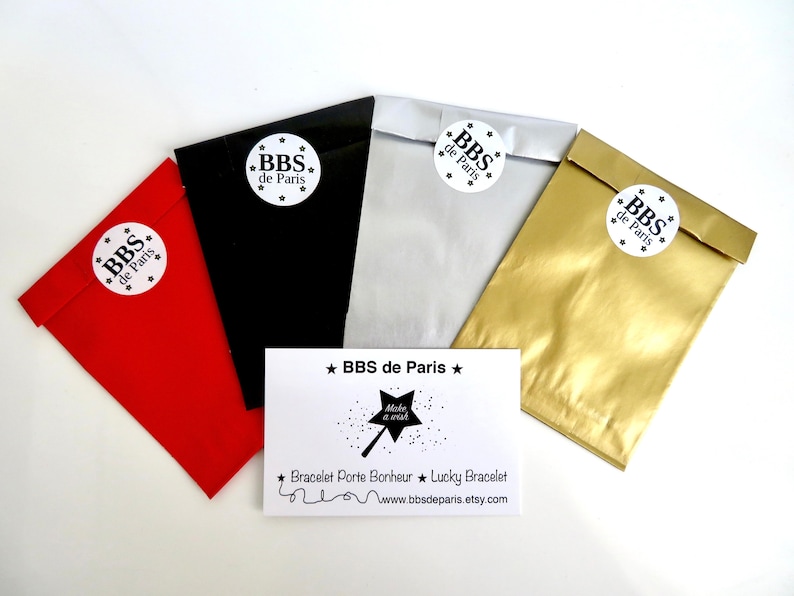 May include: Four small gift bags in red, black, silver, and gold with white circular labels that say "BBS de Paris" in black text. A white card with black text says "BBS de Paris" and "Bracelet Porte Bonheur ★ Lucky Bracelet" with a website address.