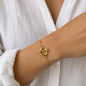 Op de afbeelding: Een goudkleurige armband met een fijne dubbele ketting en een ronde knevelsluiting. Het sieraad bestaat uit een dunne metalen ketting die verbonden is met een ronde ring en een horizontale staaf, wat een veilige knevelsluiting vormt.