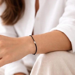 Braccialetto sottile in cordino nero con una perlina rotonda in argento sterling e due perline tubolari in argento sterling. Lettera K in codice Morse. Un portafortuna a un prezzo accessibile.