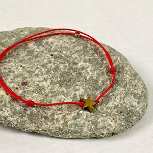 May include: A red string bracelet with a gold star charm. The bracelet is resting on a gray stone surface. The star charm is centered on the bracelet.
