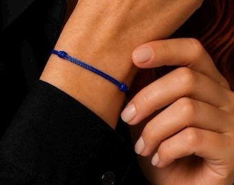 Puede incluir: Una pulsera de cordón azul real, fina y sencilla, con un diseño liso y minimalista que presenta dos pequeños nudos redondeados situados a lo largo del cordón, creando un aspecto limpio y discreto para la muñeca.