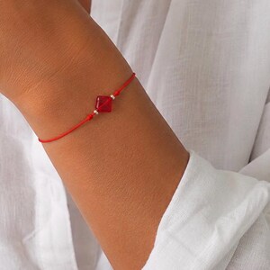 May include: A red string bracelet with a square, red gemstone and silver beads. The bracelet is worn on a wrist, against a white shirt.