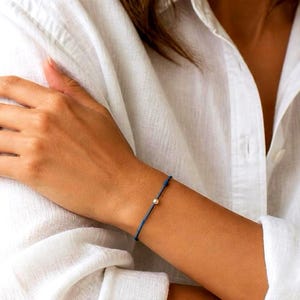 May include: A minimalist bracelet featuring a thin, dark blue cord and a single, small, round metallic bead centered on the wrist. The cord is tied with a simple knot, creating a delicate and understated accessory that rests against the skin.