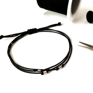 May include: Black adjustable bracelet with silver-colored beads. The bracelet is made of a thin, woven cord. Spools of black and pink thread and a pair of scissors are in the background.