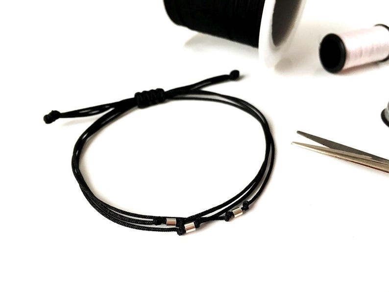 May include: Black adjustable bracelet with silver-colored beads. The bracelet is made of a thin, woven cord. Spools of black and pink thread and a pair of scissors are in the background.