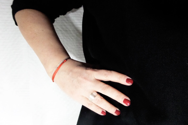 Puede incluir: Una pulsera de hilo rojo y una pulsera de plata se llevan en la mu&ntilde;eca de una persona. La persona lleva una camiseta negra y tiene esmalte de u&ntilde;as rojo en las u&ntilde;as. Un anillo de plata est&aacute; en el dedo de la persona.
