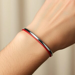 Puede incluir: Una pulsera de brazalete plateada con un fino cord&oacute;n rojo. La pulsera tiene un dise&ntilde;o simple y minimalista. El cord&oacute;n rojo es visible entre la mu&ntilde;eca y la banda plateada.