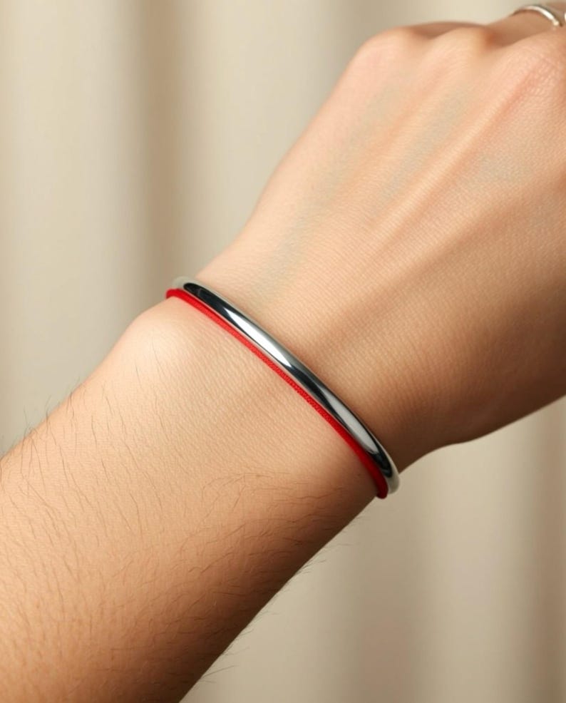 Puede incluir: Una pulsera de brazalete plateada con un fino cord&oacute;n rojo. La pulsera tiene un dise&ntilde;o simple y minimalista. El cord&oacute;n rojo es visible entre la mu&ntilde;eca y la banda plateada.