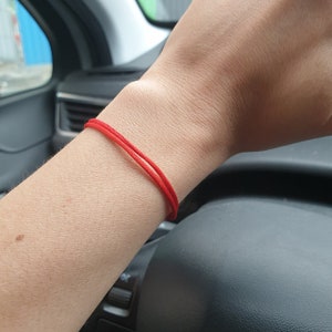 Puede incluir: Una pulsera de cuerda roja brillante, compuesta por dos cordones paralelos, se lleva en la mu&ntilde;eca. La pulsera es el punto focal, con el tono de piel de la mu&ntilde;eca y el interior de un veh&iacute;culo en el fondo.