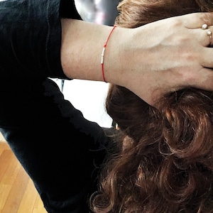 May include: A red string bracelet with a silver bar charm. The bracelet is worn on a person's wrist.
