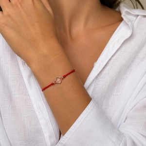 May include: A red string bracelet with a small, square, clear charm. The charm is set in a silver-colored metal. The bracelet is worn on a wrist, and the background is a white shirt.