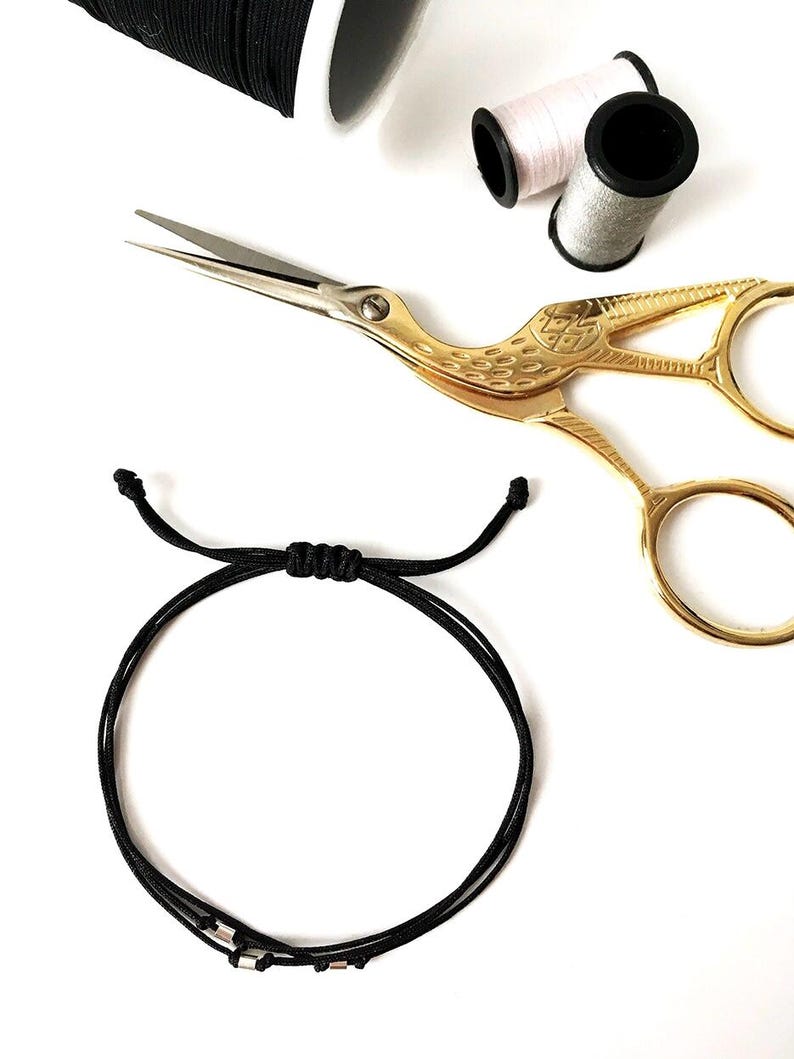 May include: A black adjustable bracelet with silver beads is displayed on a white surface. Gold scissors and spools of thread in black, pink, and gray are also visible, suggesting jewelry making or crafting.
