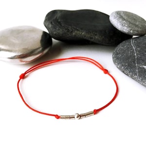 May include: A red string bracelet with silver beads. The bracelet is on a white background with gray stones.