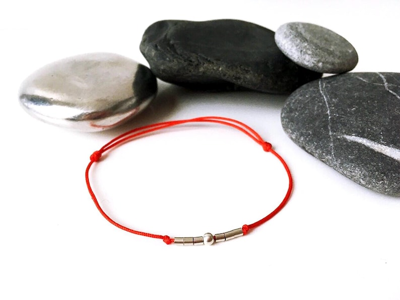 May include: A red string bracelet with silver beads. The bracelet is on a white background with gray stones.
