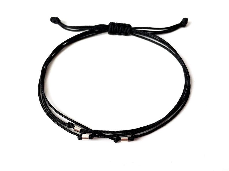 May include: Black adjustable bracelet featuring three cords and small silver-colored cylindrical beads. The bracelet is knotted at the top for adjustment and is set against a white background. A simple, minimalist design.