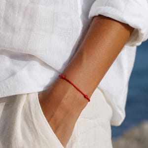 Puede incluir: Una pulsera de hilo rojo con nudos. La pulsera se lleva en la mu&ntilde;eca, sobre un fondo de ropa blanca y un fondo azul borroso.