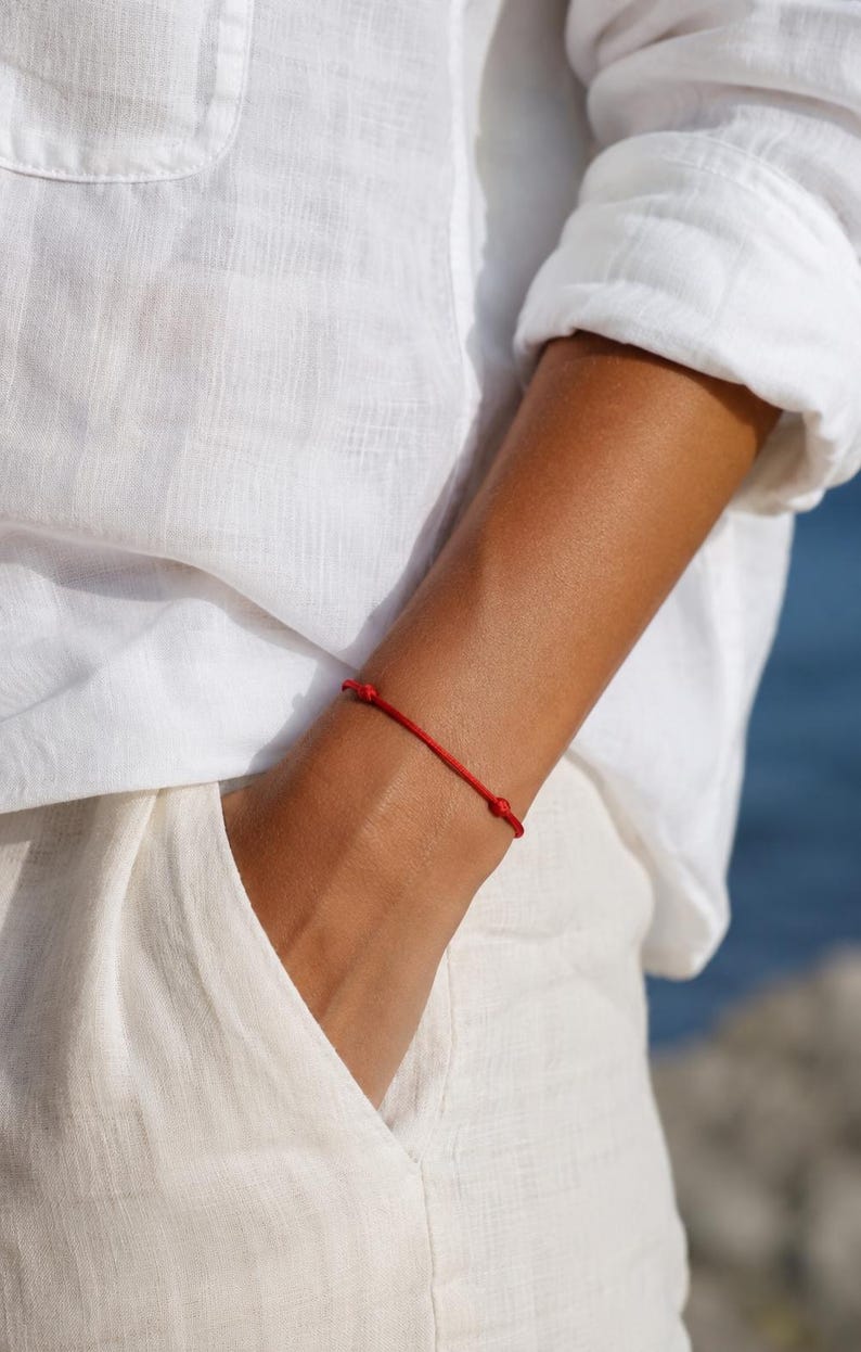 Puede incluir: Una pulsera de hilo rojo con nudos. La pulsera se lleva en la mu&ntilde;eca, sobre un fondo de ropa blanca y un fondo azul borroso.