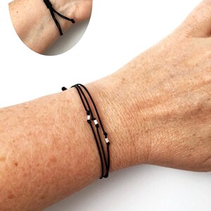 May include: A black string bracelet with three strands and small silver beads. The bracelet is tied with a sliding knot for adjustable sizing. The image shows the bracelet on a wrist, with a close-up of the knot.