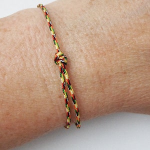 Puede incluir: Un brazalete trenzado de colores con cierre de nudo. El brazalete está hecho de hilos rojos, amarillos, verdes y negros.