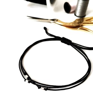 May include: A black cord bracelet with small silver-colored beads. The bracelet is displayed with sewing supplies, including spools of thread in black, pink, and gray, and gold-colored scissors, all on a white background.