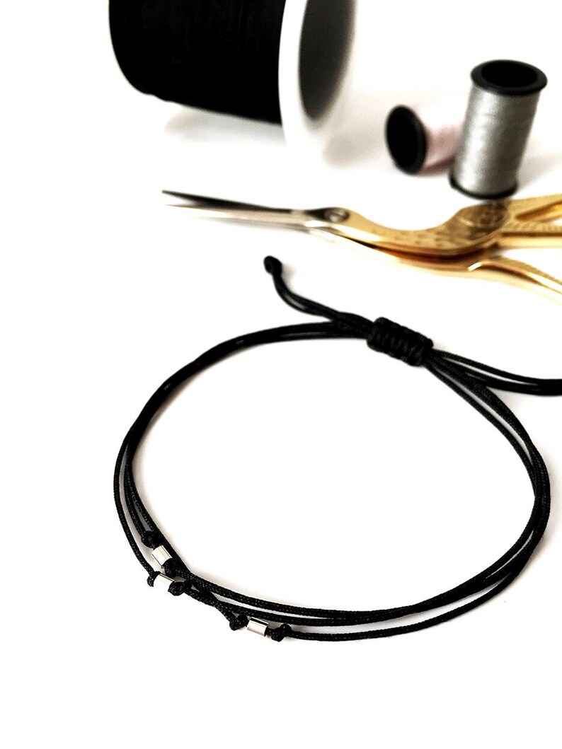 May include: A black cord bracelet with small silver-colored beads. The bracelet is displayed with sewing supplies, including spools of thread in black, pink, and gray, and gold-colored scissors, all on a white background.