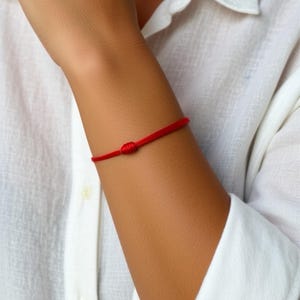 Może przedstawiać: Jasnoczerwona bransoletka z sznurka zawiązana w węzeł. Bransoletka jest noszona na nadgarstku osoby ubranej w białą koszulę. Design jest prosty i minimalistyczny.