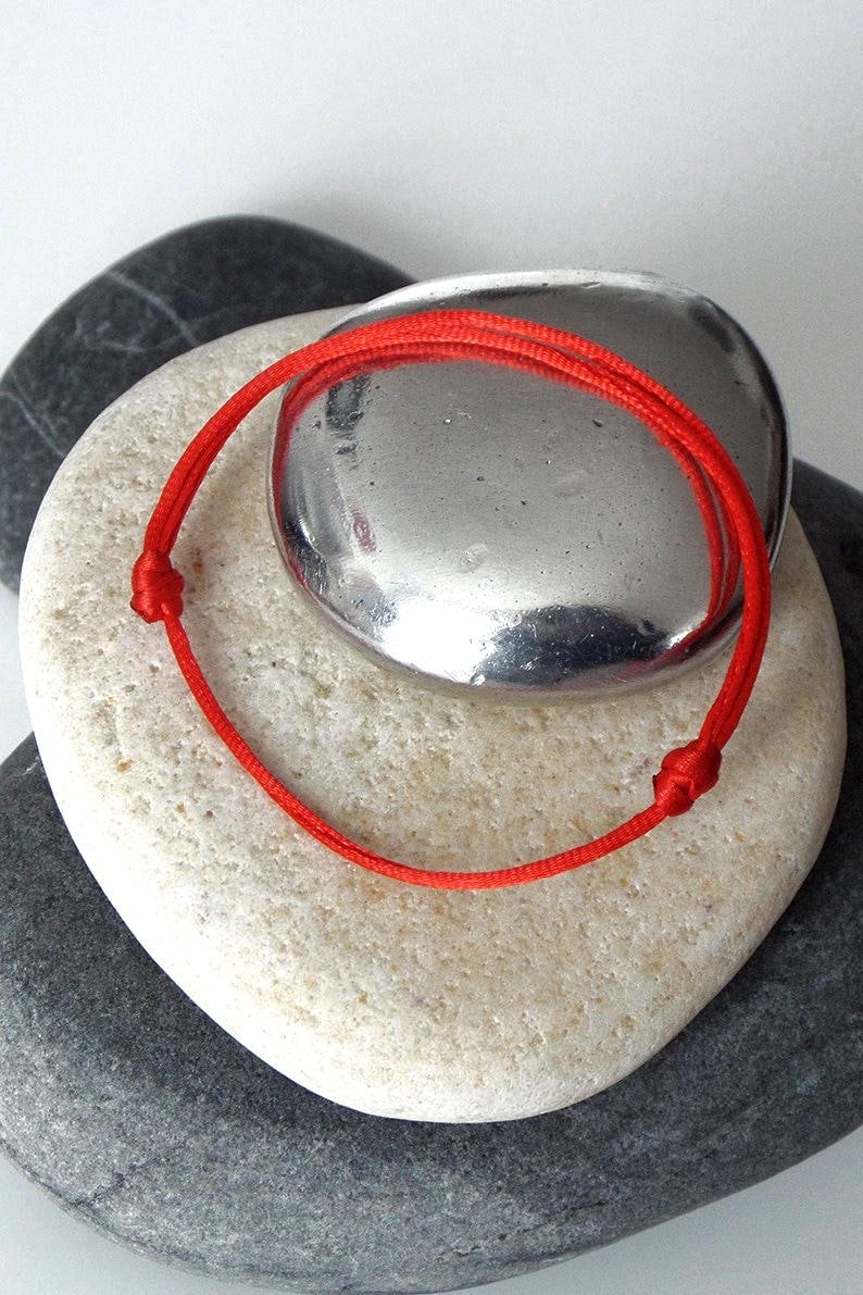 Puede incluir: Una pulsera de cord&oacute;n rojo brillante con nudos ajustables, colocada sobre una piedra lisa y clara. Tambi&eacute;n hay una piedra brillante de color plateado. La pulsera tiene un dise&ntilde;o sencillo y minimalista.