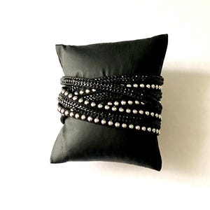 May include: A black wrap bracelet with multiple strands. The bracelet features rows of small, shiny silver beads and black chain links. The bracelet is wrapped around a small, black pillow, creating a display.