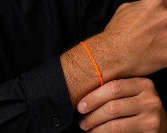 Puede incluir: Una pulsera fina de cordón en color naranja neón vibrante que presenta un mecanismo de nudo corredizo simple y ajustable. La pulsera consiste en un cordón único, liso y redondeado que rodea la muñeca, ofreciendo un accesorio minimalista y ligero.