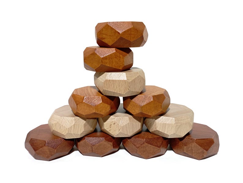 May include: A stack of wooden geometric blocks in brown and natural wood tones. The blocks are shaped like irregular pentagons and are stacked in a pyramid shape.