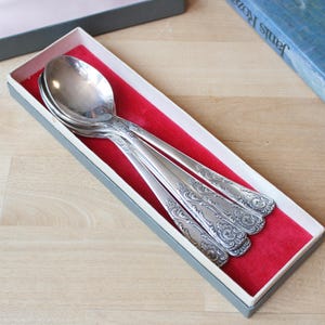Peut inclure: Un ensemble de cuillères en argent avec des motifs floraux ornés, présenté dans une boîte grise avec un intérieur en velours rouge. Les cuillères sont disposées en diagonale.