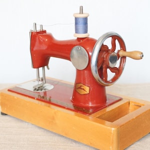Peut inclure: Une machine à coudre manuelle en métal rouge montée sur une base en bois rectangulaire. Elle est équipée d'un volant argenté avec une poignée en bois, d'un porte-bobine vertical avec un fil bleu et d'un petit logo doré sur le devant.