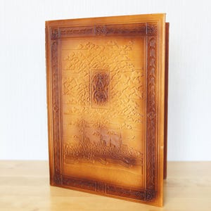 May include: A rectangular, light brown leather-bound book cover featuring an embossed, detailed scene of a cityscape with a cathedral and clouds. The cover is framed by a decorative border with intricate patterns and a central rectangular emblem.