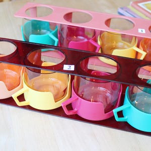 Peut inclure: Ensemble de tasses en verre vintage colorées, chacune avec une poignée et une couleur différente, rose, jaune, orange et turquoise. Les tasses sont maintenues dans un support en plastique rouge foncé avec des ouvertures circulaires.