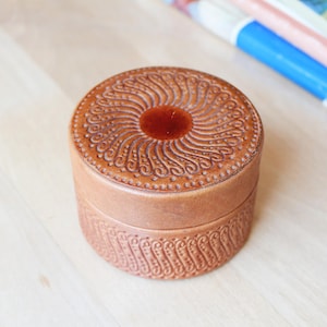 Könnte beinhalten: Eine runde, hellbraune Lederdose mit einer glatten, kreisförmigen Mitte auf dem Deckel, die von einem komplexen, geprägten Spiralmuster umgeben ist. Die Seiten der Dose zeigen zudem ein sich wiederholendes, geprägtes vertikales Wellenmuster.