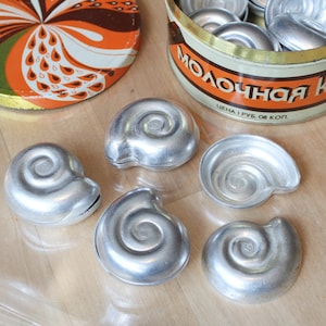 May include: A collection of vintage, silver-colored shell-shaped molds, likely for baking. They are arranged on a light-colored surface, alongside a tin container filled with more molds and a patterned lid.