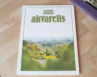 May include: A book titled "Latviešu Mūsdienu Akvarelis" with a landscape painting on the cover. The cover art features green and yellow foliage with a misty background. The book is on a light-colored wooden surface.